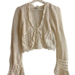 Vit stickad cardigan - Charmig vit stickad cardigan/kofta med vackra spetsdetaljer och knytning framtill. Koftan har långa vida ärmar och en croppad passform, perfekt för lager-på-lager-stil. Den är både söt och stilren, vilket gör den till ett mångsidigt tillskott i garderoben. Helt oanvänd från Urban Outfitters. Storlek L men passar även S-M