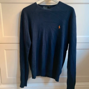 Mörkblå tröja från Polo Ralph Lauren - Säljer en stilren mörkblå tröja från Polo Ralph Lauren i storlek M. Tröjan är tillverkad av 100% bomull och har en slim fit passform. Den har långa ärmar och en liten broderad logotyp i orange på bröstet. Perfekt för en klassisk look.