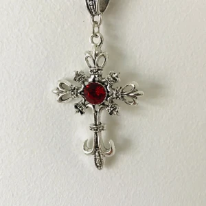 Silverfärgat korshalsband med röd sten - Elegant silverfärgat korshalsband med en stor röd sten i mitten. Korset har detaljerade utsmyckningar och hänger från en dekorativ kedja. Perfekt för att ge en stilfull touch till din outfit.