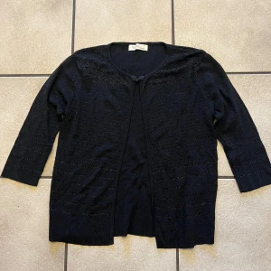 Svart kofta  - Elegant svart kofta med trekvartsärmar och subtila glittriga detaljer. Perfekt för att lägga till en touch av stil till din outfit. Koftan har en enkel knäppning framtill.