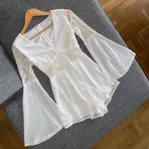 Vit spets playsuit från Rare London, storlek XS - Snygg vit playsuit från Rare London med spetsdetaljer och vida ärmar. Den har en v-ringad design och dragkedja bak. Perfekt för en stilren look.
