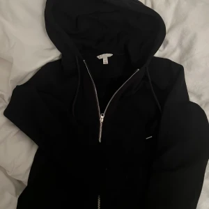 Svart hoodie med dragkedja från H&M - Säljer en svart hoodie från H&M med dragkedja och huva. Den har en klassisk design med snören vid huvan och är tillverkad i ett mjukt material. Säljer pga av att den aldrig kommit till användning.