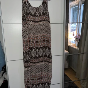 Mönstrad långklänning från Indiska - Säljer en snygg långklänning från Indiska med ett unikt mönster i brunt, orange och vitt. Klänningen har en ärmlös design och är perfekt för en bohemisk stil. Den är tillverkad i ett mjukt och bekvämt material.