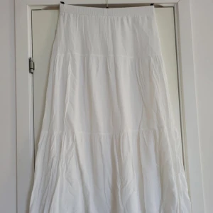 Vit långkjol - Säljer en vacker vit långkjol med ett luftigt och flödande tyg. Kjolen har en enkel och elegant design som passar perfekt för varma dagar. Den är gjord i ett lätt material som ger en skön känsla.