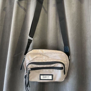 Beige axelväska  - Snygg beige liten axelväska från DAY ET med justerbar svart axelrem. Väskan har flera dragkedjeförsedda fack för praktisk förvaring. Perfekt för att bära med sig det nödvändigaste på ett stilrent sätt.