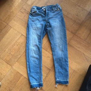 Blå wide jeans från Lindex, storlek 146/10-11 år - Säljer ett par blå jeans från Lindex med en avslappnad wide fit. Byxorna har slitna detaljer vid bensluten för en trendig look. Perfekta för en casual stil.