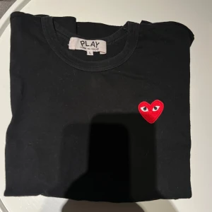 Svart t-shirt från Comme des Garçons - Säljer en svart t-shirt från Comme des Garçons med det ikoniska röda hjärtat broderat på bröstet. Perfekt för en stilren och trendig look. T-shirten har en klassisk passform och är tillverkad i mjuk bomull. Den är använd fåtal gånger och är i L men skulle säga att den är som en S i storlek!
