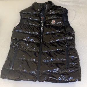 Svart glansig dunväst från Moncler - Snygg svart glansig dunväst från Moncler med dragkedja och broderad logga på bröstet. Perfekt för kyliga dagar med sin quiltade design som håller värmen. Västen har en hög krage och två praktiska fickor med dragkedja. Den passar bra till storlek s och m