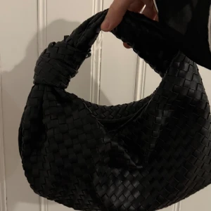 Svart flätad handväska - Snygg svart handväska med flätat mönster och knutdetalj på handtaget. Perfekt för att ge en stilren touch till din outfit. Väskan är kompakt och lätt att bära med sig.