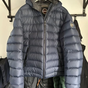 Blå pufferjacka från Colmar - Säljer en snygg blå pufferjacka från Colmar. Jackan har en quiltad design med dragkedja framtill och en huva för extra värme. Perfekt för kyliga dagar!