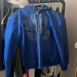 Blå rhinestone hoodie från Ahead Of Time - Säljer en blå huvtröja från Ahead Of Time med dragkedja och glittriga detaljer på framsidan. Tröjan har långa ärmar och en bekväm passform. Perfekt för en avslappnad stil.