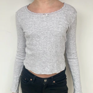 Grå långärmad topp från H&M - Säljer en söt grå långärmad topp från H&M med spetsdetaljer och en liten rosett vid halsringningen. Toppen har ett diskret mönster och är croppad. Den kommer inte till användning längre och den är bra skick och väldigt bekväm.