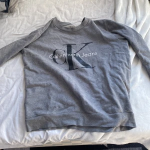 Grå sweatshirt från Calvin Klein Jeans - Säljer en stilren grå sweatshirt från Calvin Klein Jeans med stort CK-logotyp tryck på framsidan. Tröjan har långa ärmar och en bekväm passform, perfekt för en avslappnad look. Passar bra till jeans eller joggers. 🖤