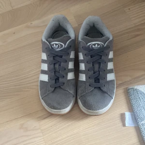 Grå Adidas Campus sneakers - Snygga grå Adidas Campus sneakers i mocka med klassiska vita ränder på sidorna och vit logga på hälen. Skorna har rund tå, platt sula och snörning. Perfekta för dig som gillar en clean och tidlös streetstil.