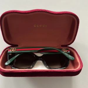 Gucci solglasögon med gröna skalmar - Snygga solglasögon från Gucci med markerad fyrkantig form. Skalmarna är gröna med röd rand och guldigt GG-logo på sidorna. Framsidan är mörkbrun med svarta detaljer och glasen är mörkt tonade. Kommer med originalfodral och röd silkespåse. Självklart äkta, kvitto finns kvar, köpta för 2800 2023 och har varit extremt sparsamt använda! 