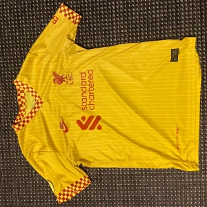 Liverpool Salah bortatröja Nike gul - Liverpool FC bortaställ med M. Salah 11 på ryggen. Tröjan är gul med tunna vita ränder, röda tryck och schackrutiga detaljer i rött och gult på ärmslut och krage. Nike Dri-FIT-material som andas. Matchande shorts medföljer.