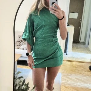 Grön one shoulder kortklänning - Snygg grön kortklänning med one shoulder-design och en ärm. Klänningen har draperad midja och knyt-detalj på.