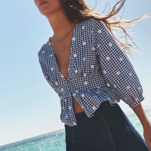 Blåvit rutig blus från Zara - Supersöt blus från Zara i blåvitt rutigt mönster med små broderade vita blommor. Djup v-ringning, knappar framtill och lätt puffade långa ärmar. Modellen är croppad med volang nedtill och sydd i luftig bomull. Perfekt för sommarens alla äventyr!