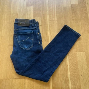 Jacob Cohën mörkblå jeans slim fit - Mörkblå jeans från Jacob Cohën med klassisk femficksdesign, snygga kontrastsömmar och läderpatch bak i midjan. Jeansen har en slim passform och är tillverkade i mjukt bomullsjeansmaterial med lite stretch för extra komfort.