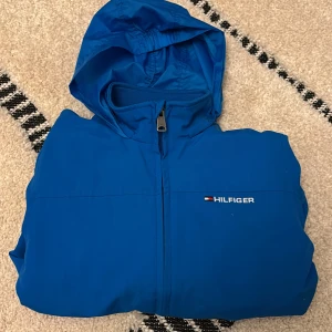 Blå vindjacka från Tommy Hilfiger - Snygg blå vindjacka från Tommy Hilfiger med huva och dragkedja framtill. Jackan har logga på bröstet och ärmen, samt meshfoder på insidan för extra komfort. Perfekt för blåsiga dagar och enkel att matcha med streetwear.