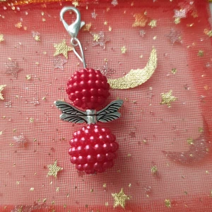 Röd hallon-berlock med silvervingar - Unik berlock med två röda hallonformade pärlor och detaljerade silverfärgade vingar mellan dem. Perfekt att hänga på halsband eller armband för en söt och lekfull look. Fästet är i silverfärg och gör det enkelt att sätta fast på din favoritkedja. Handgjord smyckesängel 