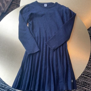 Mörkblå glittrig plisserad klänning från petit bateau - Supersnygg mörkblå klänning med glittriga trådar och plisserad kjol. Långärmad modell med rund halsringning och mjukt stickat material. Perfekt för dig som vill sticka ut lite extra med en klassisk men ändå trendig look.