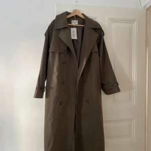 Brun trenchcoat från NA-KD - Säljer en brun trenchcoat från NA-KD med klassisk dubbelknäppning, bred krage och långa ärmar med justerbara remmar vid handlederna. Oversized och är perfekt för lager-på-lager. Ny med prislapp 