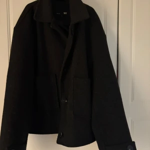Svart kappa  från 157 - Snygg svart jacka från 157 i ull med klassisk krage och stora knappar framtill. Jackan har två rejäla fickor och en rak, boxig passform som ger en chill vibe. Perfekt för lager-på-lager och funkar till allt när det är kallt ute.