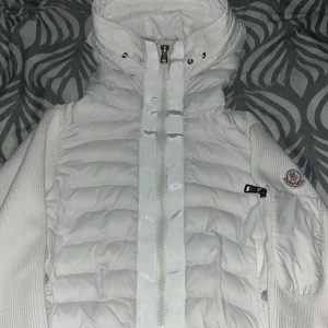 Vit Moncler pufferjacka med huva - Snygg vit pufferjacka från Moncler med huva och dragkedja framtill. Jackan har ribbade stickade ärmar och Moncler-logga på ena ärmen. Fodret är quiltat och jackan har en cool serietidningsdetalj på insidan. Perfekt för dig som vill ha en clean och exklusiv look.  