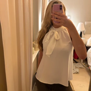 Vit ärmlös blus med knyt i halsen - Säljer en vit, ärmlös blus med hög hals och snygg knytrosett framtill. Aldrig använd så därav i nyskick! Storlek S men passar även M  