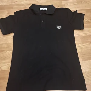 Svart Stone Island pikétröja - Svart piké från Stone Island med klassisk krage och knappslå framtill. T-shirten har korta ärmar och en snygg broderad logotyp på bröstet. Materialet är bomull och passformen är normal, perfekt för en clean och stilren look.