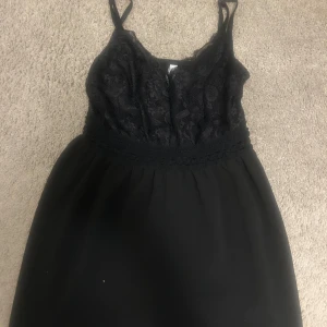 Svart spetsklänning med smala axelband - Snygg svart kortklänning med smala axelband. Klänningen har inbyggda shorts  i kjolen och ett markerat midjeband med spetsdetaljer. Perfekt för dig som gillar en stilren look med lite extra detaljer.