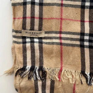  Burberry halsduk - Klassisk halsduk från Burberry London med ikoniskt rutigt mönster i beige, svart, vitt och röda linjer. Halsduken har fransar i kanterna och känns både tidlös och trendig. Perfekt accessoar för att lyfta din outfit.