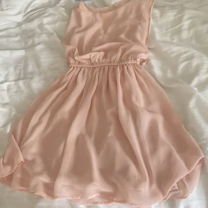 Ljusrosa klänning med pärldetaljer - Supersöt ljusrosa klänning med pärlor längs ärmhålen. Den har ett mjukt, flowy tyg och resår i midjan som ger en snygg siluett. Klänningen är ärmlös och har en knapp i nacken. 