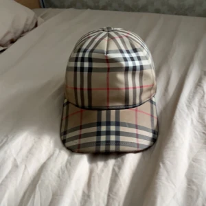 Äkta, Burberry rutig keps i beige och svart - Snygg keps från Burberry med klassiskt rutigt mönster i beige, svart, vitt och röda linjer. Justerbar baktill med svart spänne och tydlig Burberry London England-logga. Perfekt accessoar för att lyfta din outfit.