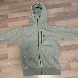 Grå hoodie från Sail Racing XS - Snygg grå hoodie från Sail Racing i storlek XS. Tröjan har huva med snörning, diagonal dragkedja och en extra zipficka på bröstet med logga. Mjuk insida i fleece och långa ärmar. Perfekt för chill eller när du vill vara bekväm.