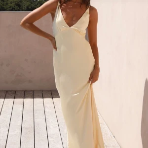 Champagnefärgad långklänning med v-ringning - Säljer en elegant långklänning i champagnefärg med djup v-ringning och smala axelband. Klänningen har en figurnära passform och är tillverkad i ett glansigt, mjukt material som ger en lyxig känsla. Perfekt för student eller bal! Skriv vid frågor.