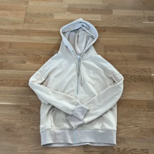 Beige hoodie från Ami med dragkedja - Säljer en beige hoodie från Ami med dragkedja och huva. Tröjan har ett broderat hjärta med bokstaven A på bröstet, ribbade muddar och fickor framtill. Perfekt för chill dagar och snygg att matcha med jeans eller joggers. Står Xl men skulle säga att den är mer som L