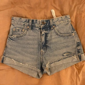 Blå jeansshorts från Pull&Bear - Snygga blå jeansshorts från Pull&Bear med hög midja och råa, fransiga kanter. De har slitna detaljer både fram och bak, klassiska fem fickor och knappstängning. Perfekta för sommarens chill dagar och festivalhäng.