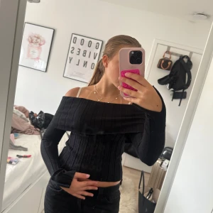 Svart ribbad offshoulder korsettopp - Säljer en svart ribbad offshoulder korsettopp med långa ärmar och croppad passform. Toppen har en snygg figurnära siluett och bredare kant upptill som ger en cool look. Perfekt att styla med jeans eller kjol för en trendig vibe.