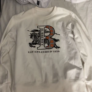 Vit sweatshirt från Burberry - Säljer en vit sweatshirt från Burberry med stort B-tryck och klassiskt rutig mönster i orange och svart på bröstet. Tröjan har rund hals och långa ärmar, perfekt för dig som gillar stilrena designer med en twist.