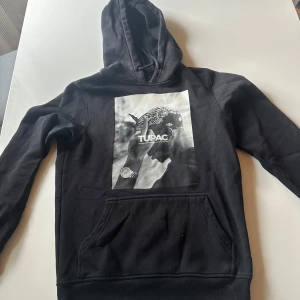 Svart Tupac hoodie från Mister Tee XS - Svart hoodie från Mister Tee i storlek XS med stor Tupac-print på bröstet. Tröjan har huva med snörning och en klassisk magficka. Perfekt för dig som gillar streetstyle och vill ha en cool look med ikonisk hiphop-vibe.