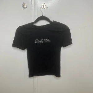 Svart croppad topp från Bikbok XS - Svart croppad topp från Bikbok i storlek XS med korta ärmar. På bröstet står det 'Dolce Vita' i glittriga stenar som ger en extra touch. T-shirten har en slim passform och är gjord i mjuk bomull. Perfekt för dig som gillar stilrena plagg med lite bling.