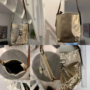 Guldig axelväska med tofsdetalj - Ny Snygg axelväska i guldigt syntet med coola utskurna mönster och en stor tofs framtill. Väskan har brunt axelband och dragkedja upptill. Ca 26cm hög utan axelbandet. Hämtas i Hägersten 