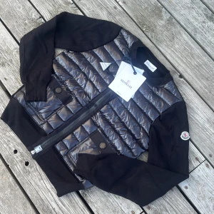  Moncler cardigan - Snygg svart pufferjacka från Moncler med glansig quiltad front och rygg, samt stickade ärmar och muddar. Jackan har två fickor med knapp framtill, dragkedja och klassisk Moncler-logga på ärmen. Perfekt för dig som vill ha en stilren och exklusiv look. Storlek 1
