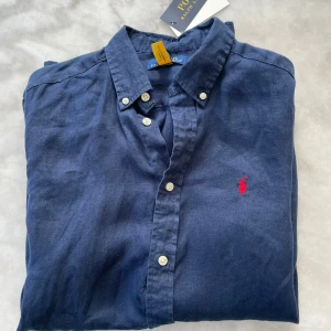 Polo Ralph Lauren skjorta - Snygg marinblå skjorta från Polo Ralph Lauren i linne. Klassisk button-down med vita knappar och röd broderad logga på bröstet. Modellen är 173 och sitter lite långt på han. Perfekt för en clean och avslappnad look. Vid minsta beundran hesitera inte att fråga.