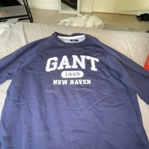 Blå sweatshirt från GANT med tryck - Säljer en marinblå sweatshirt från GANT i storlek M. Tröjan har ett stort vitt tryck med texten 'GANT 1949 NEW HAVEN' på bröstet och rund hals. Perfekt för dig som gillar klassisk college-stil och vill ha en skön tröja i bomull.