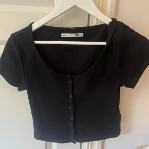 Svart croppad topp med knappar - Svart croppad topp från 157 med korta ärmar och knappar hela vägen framtill. Toppen har en rundad urringning och är gjord i ett ribbat material som ger en snygg och bekväm passform. Perfekt att styla med höga jeans eller kjol.