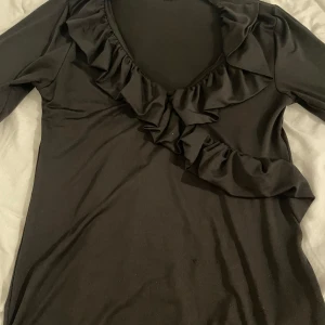 Svart volangblus från Amisu - Svart långärmad blus från Amisu med snygg v-ringning och volangdetaljer längs halsringningen. Toppens material är mjukt och stretchigt, perfekt för dig som gillar en stilren look med lite extra edge. Passar dig som vill ha något enkelt men ändå med en twist.