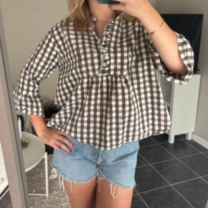 Brun och vit rutig blus med volang - Superfin blus i brun och vit rutig bomull med volangdetalj längs knäppningen framtill. Blusen har trekvartsärm och en loose passform som ger en chill vibe. Köpt på Vinted, i bra skick! Bilden är lånad av förra säljaren